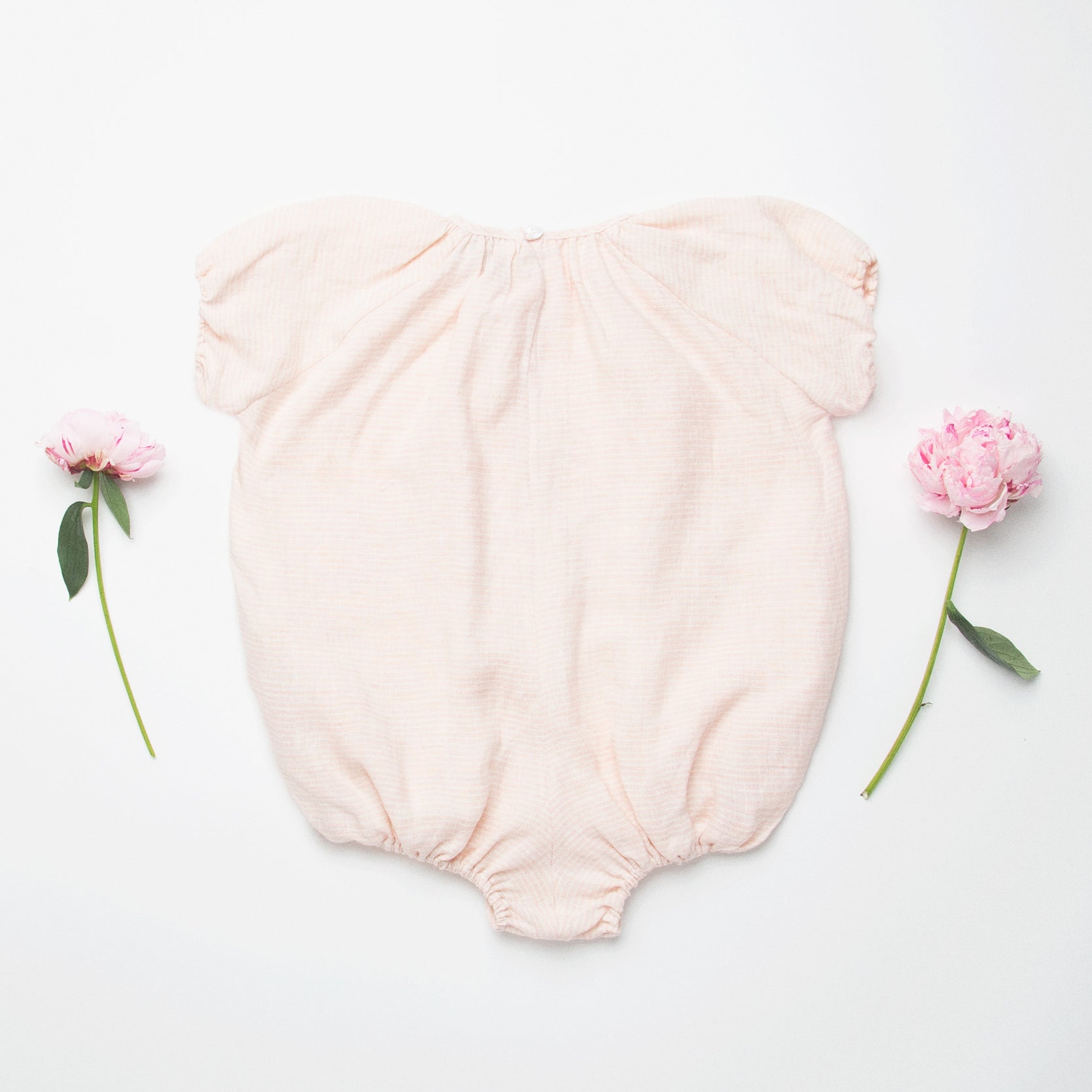 All in one romper, with short puffed elasticated sleeves and gathered neck to create a full, loose body shape. Closes at the back with single mother of pearl button, has elasticated leg holes and poppers for ease of opening. Designed to sit at the top of the thigh, perfect layered over tights in cooler weather and without in the summer. Made from super soft pale pink stripe linen.